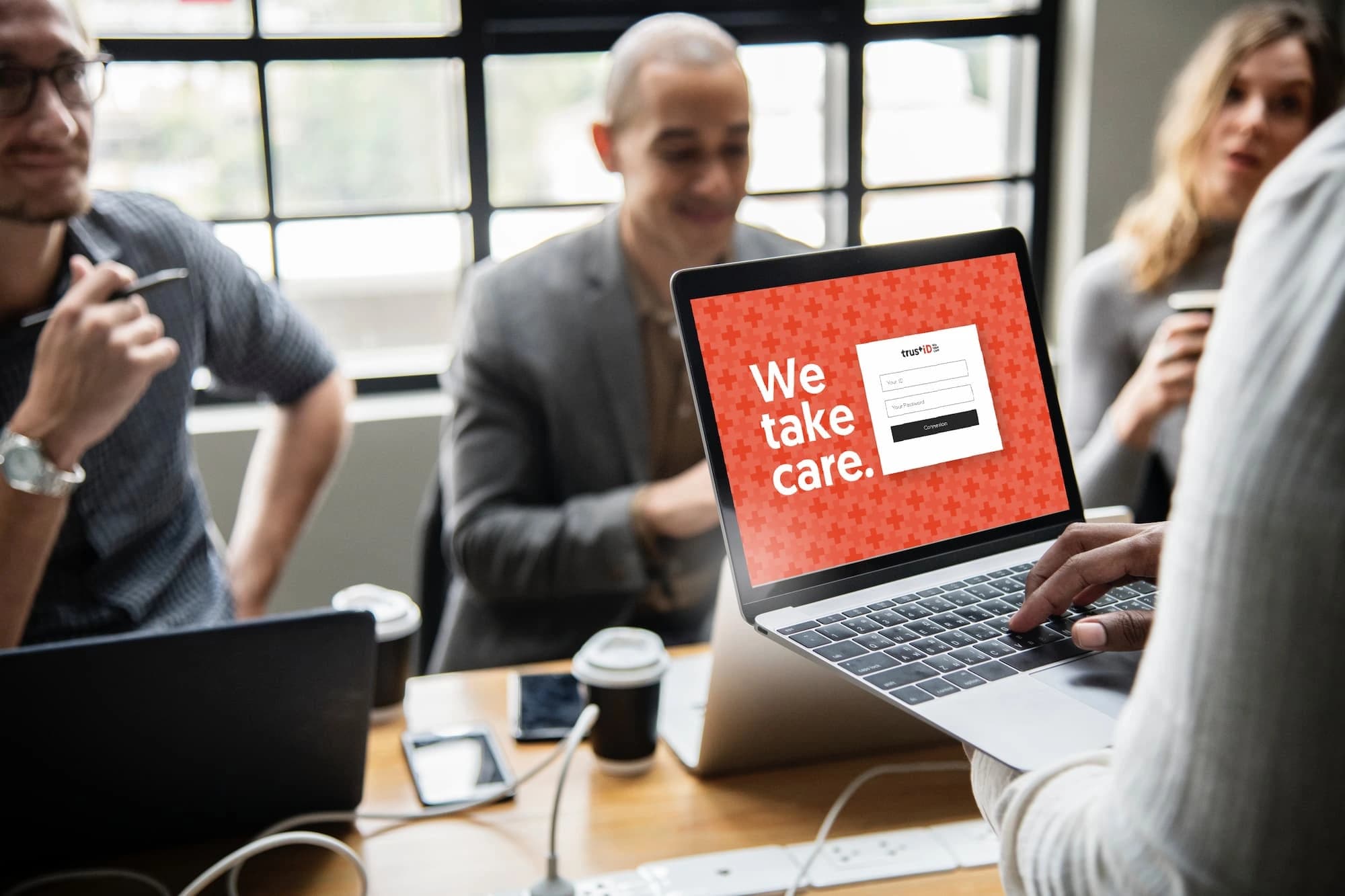 Person holding a laptop displaying a TrustID login screen with the message 'We take care' during a business meeting with three colleagues sitting around a table, coffee cups and electronic devices visible